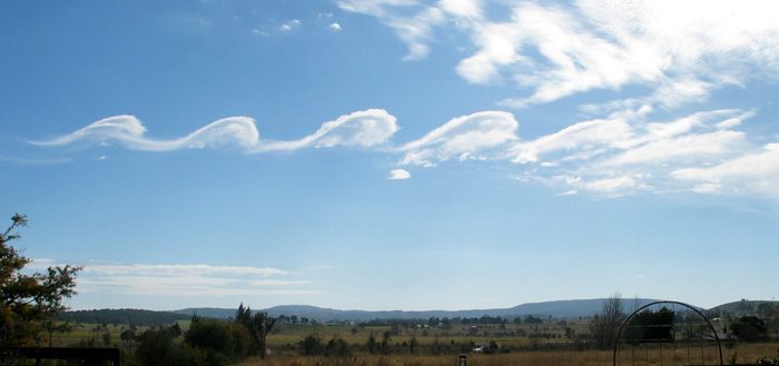 A row of wave-shaped cloud crests curling over along the top of a cloud layer above hills.