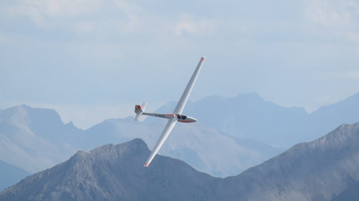 A white glider banked steeply over hazy mountains, seen from above so its very long, narrow wings stand out.
