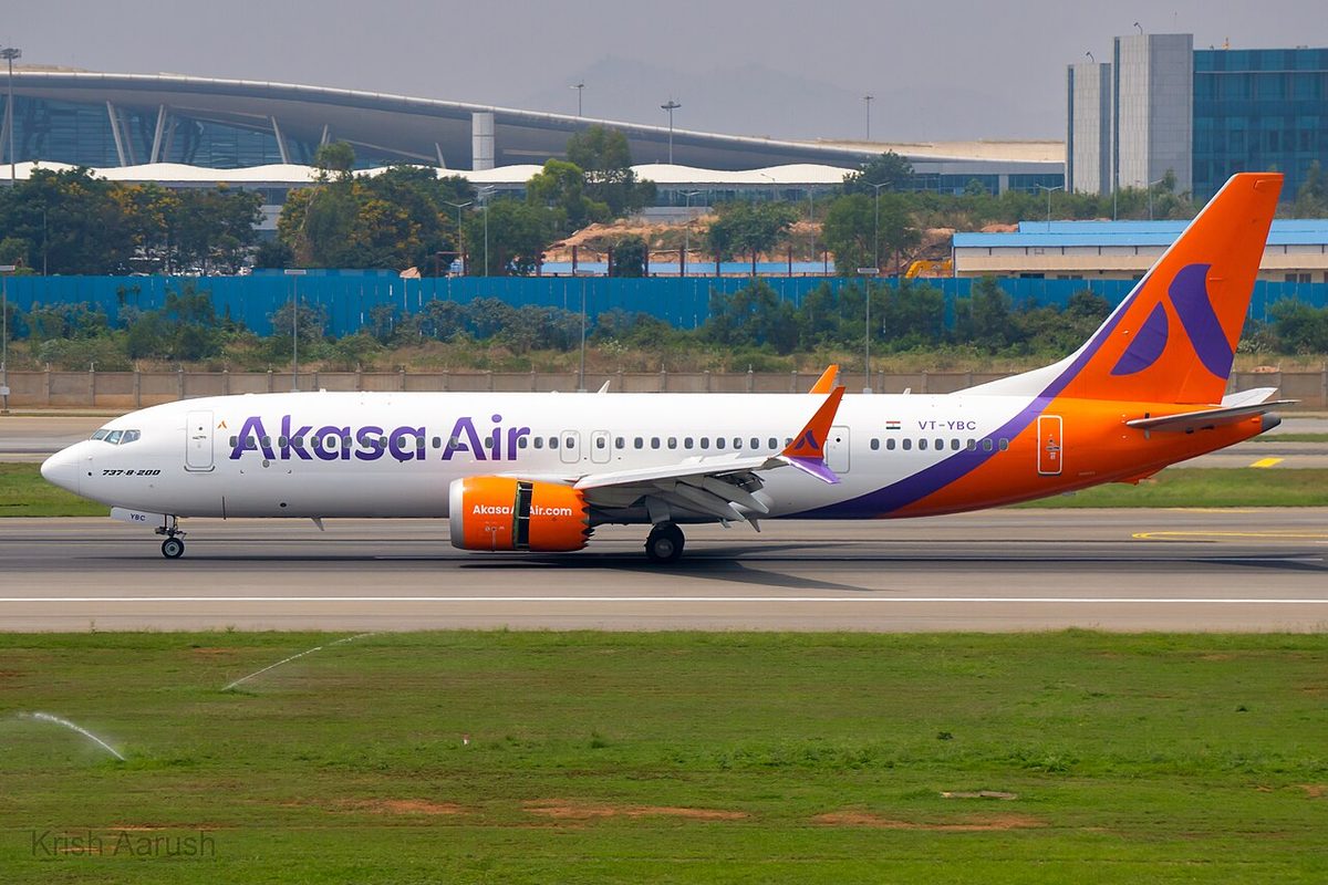 A white Akasa Air Boeing 737 MAX with an orange tail and orange engines on the runway at Bengaluru, 737-8-200 written on its nose, grass in the foreground.
