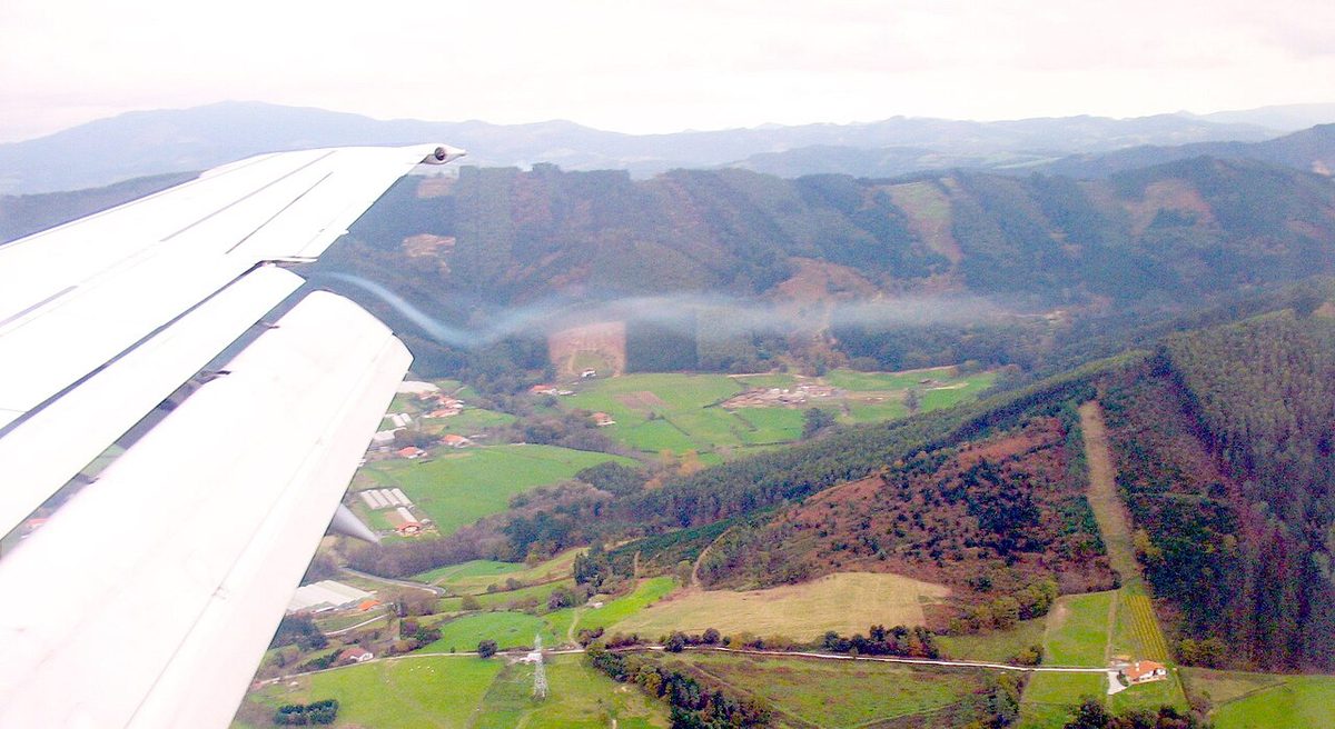 View from a cabin window of an airliner wing with flaps extended, and a thin trail of condensation spiralling back from the wingtip over hilly countryside.