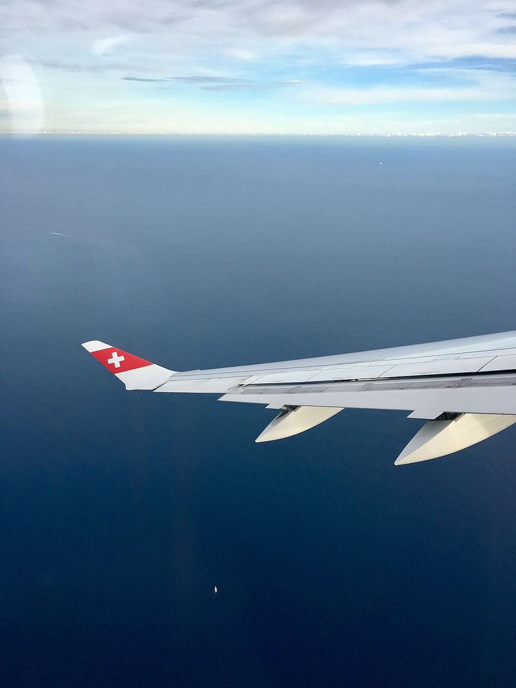 View from a cabin window of an airliner's swept wing over the sea, the wingtip in red and white with a Swiss cross, two streamlined fairings under the trailing edge.