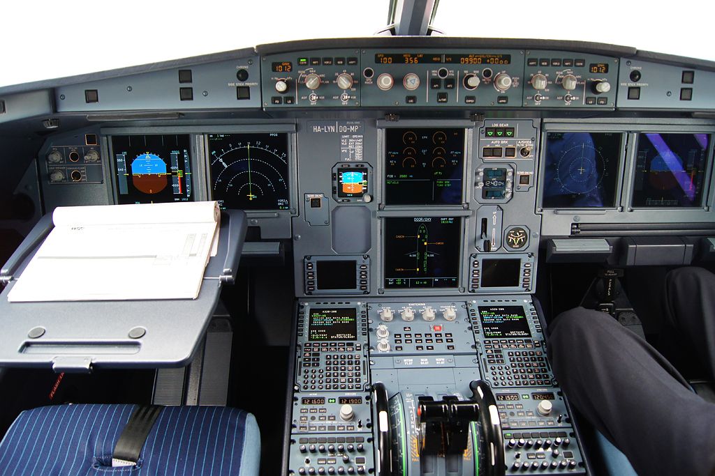 Airbus flight deck seen from behind the seats: two screens stacked in the centre panel, pilot's flight displays on the left and pedestal below.