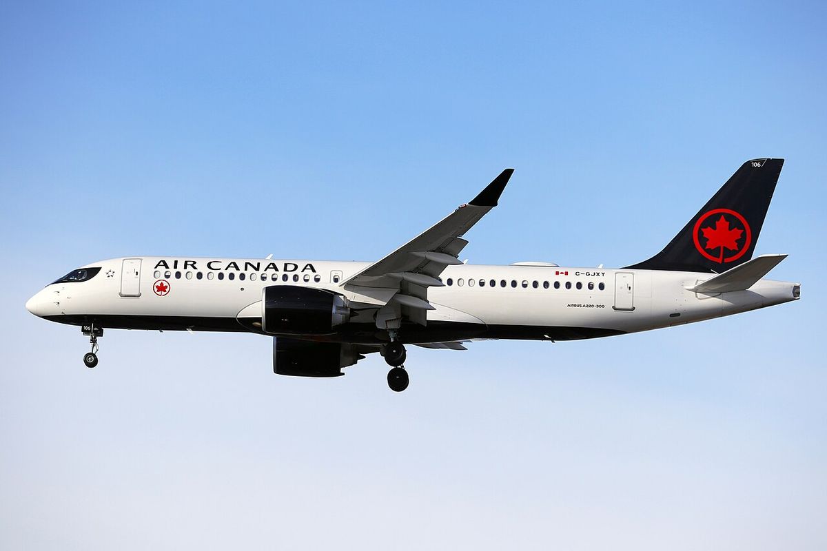 A white Air Canada Airbus A220 with a black belly and a black tail with a red maple leaf, landing gear down, against a pale sky.