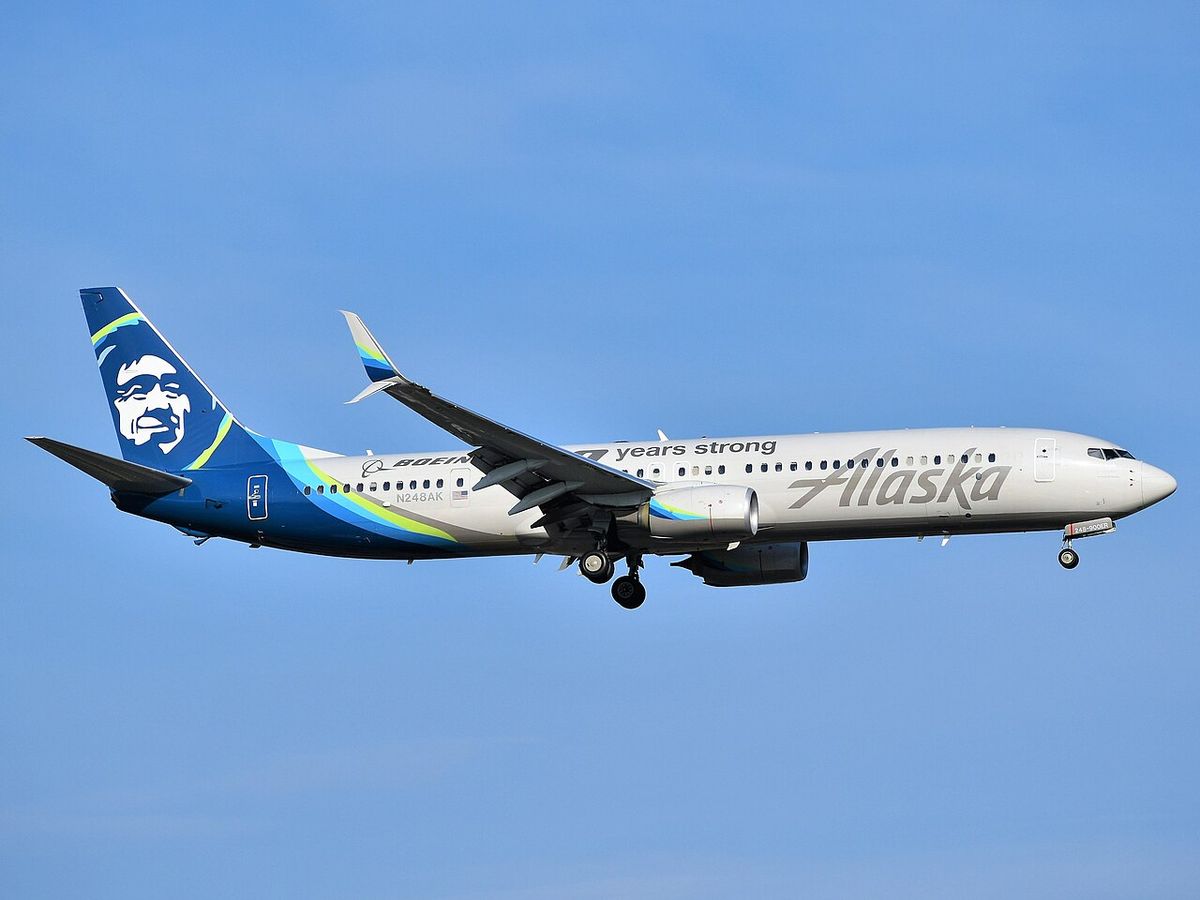 An Alaska Airlines Boeing 737-900ER on final approach with its landing gear down, in the airline’s blue and teal livery.