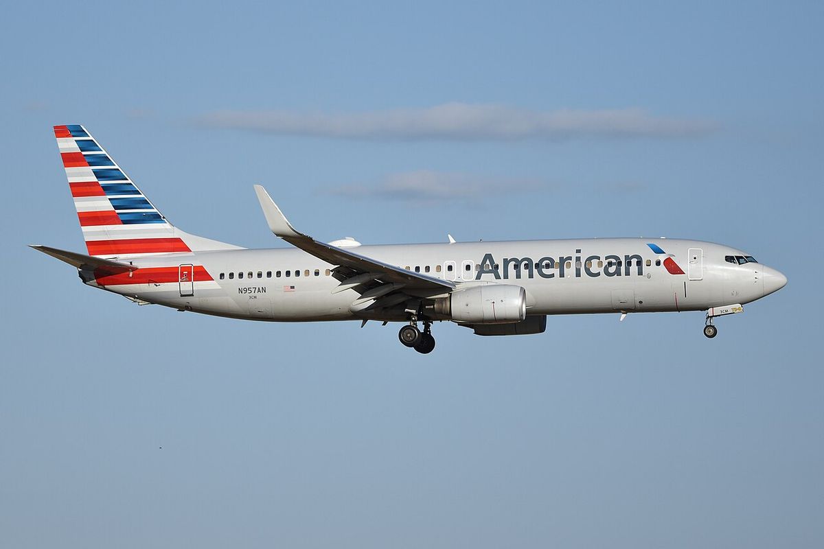 A silver American Airlines Boeing 737 with red, white and blue tail stripes flying with landing gear down.