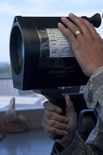 Close view of a large black signal light gun held up in both hands at a control tower window.