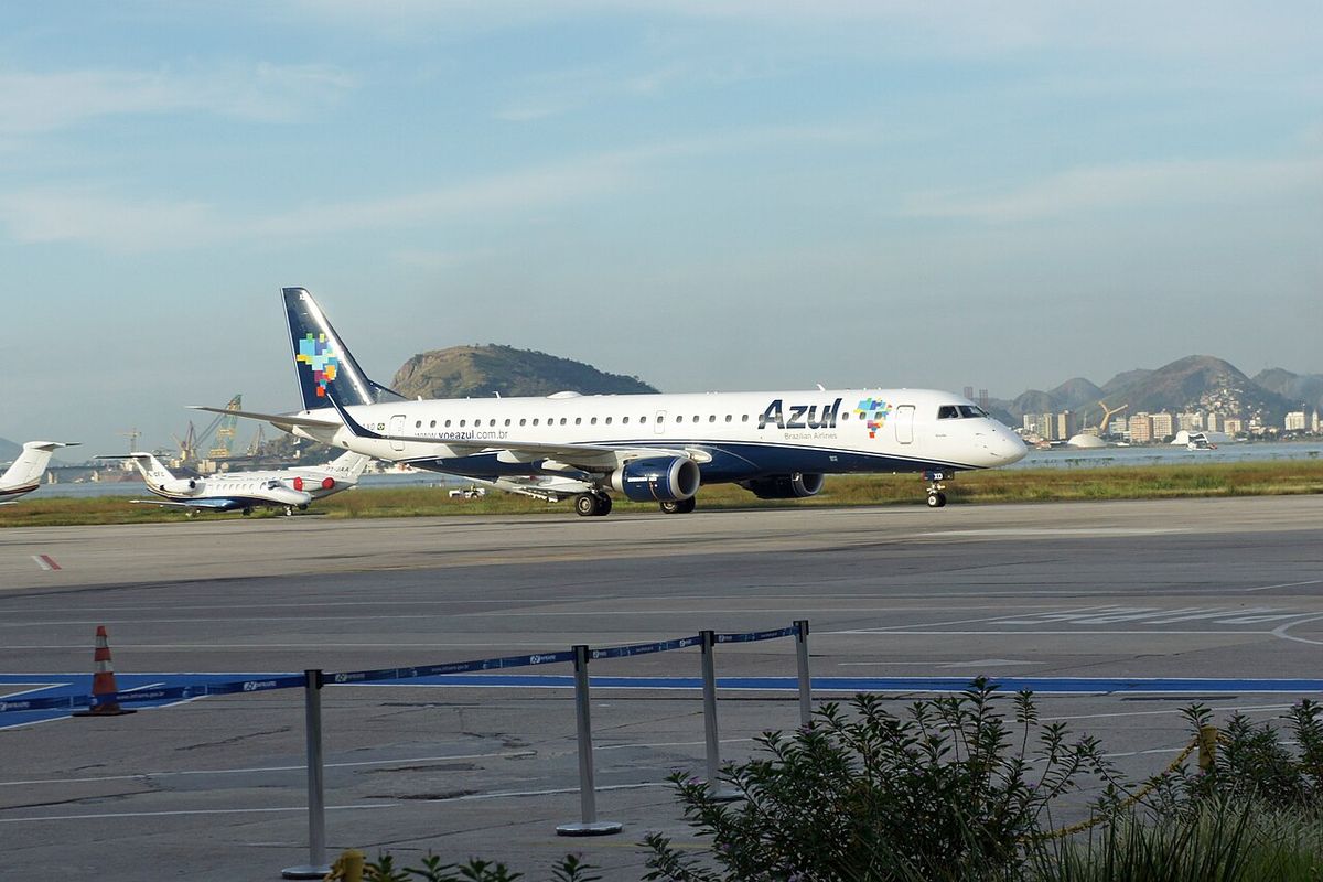 A white Azul Embraer 195 regional jet with a dark blue tail taxiing, mountains and parked aircraft behind a fence.
