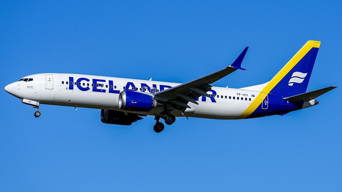 White airliner with blue lettering and a blue and yellow tail flying against a clear blue sky, landing gear down.