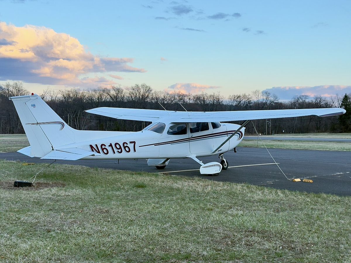 A white high-wing Cessna 172 registered N61967 parked on grass beside a paved apron under a blue evening sky.