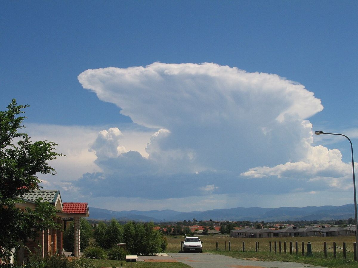 A towering white storm cloud with a flat anvil top spreading out to the left over clear sky, above a rural road, fence and distant hills.