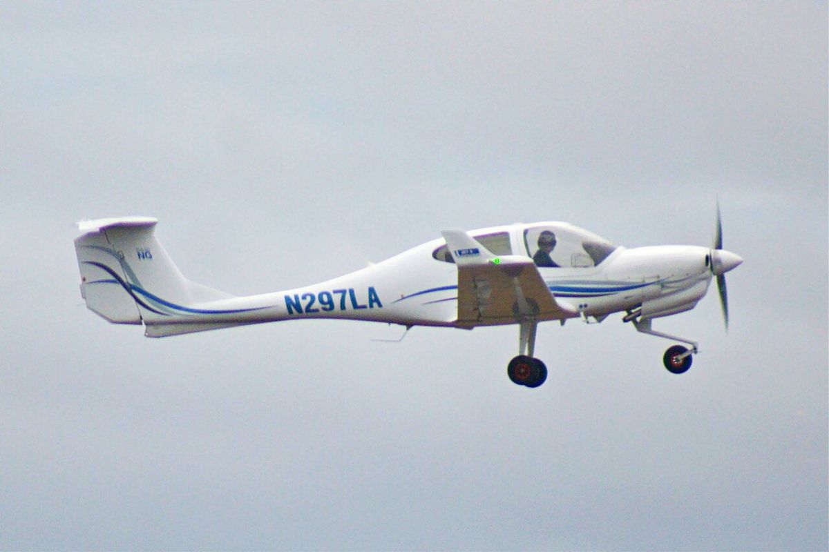 A white single-engine Diamond DA40 with a T-tail and N297LA on the side flying low with its gear down against a grey sky.