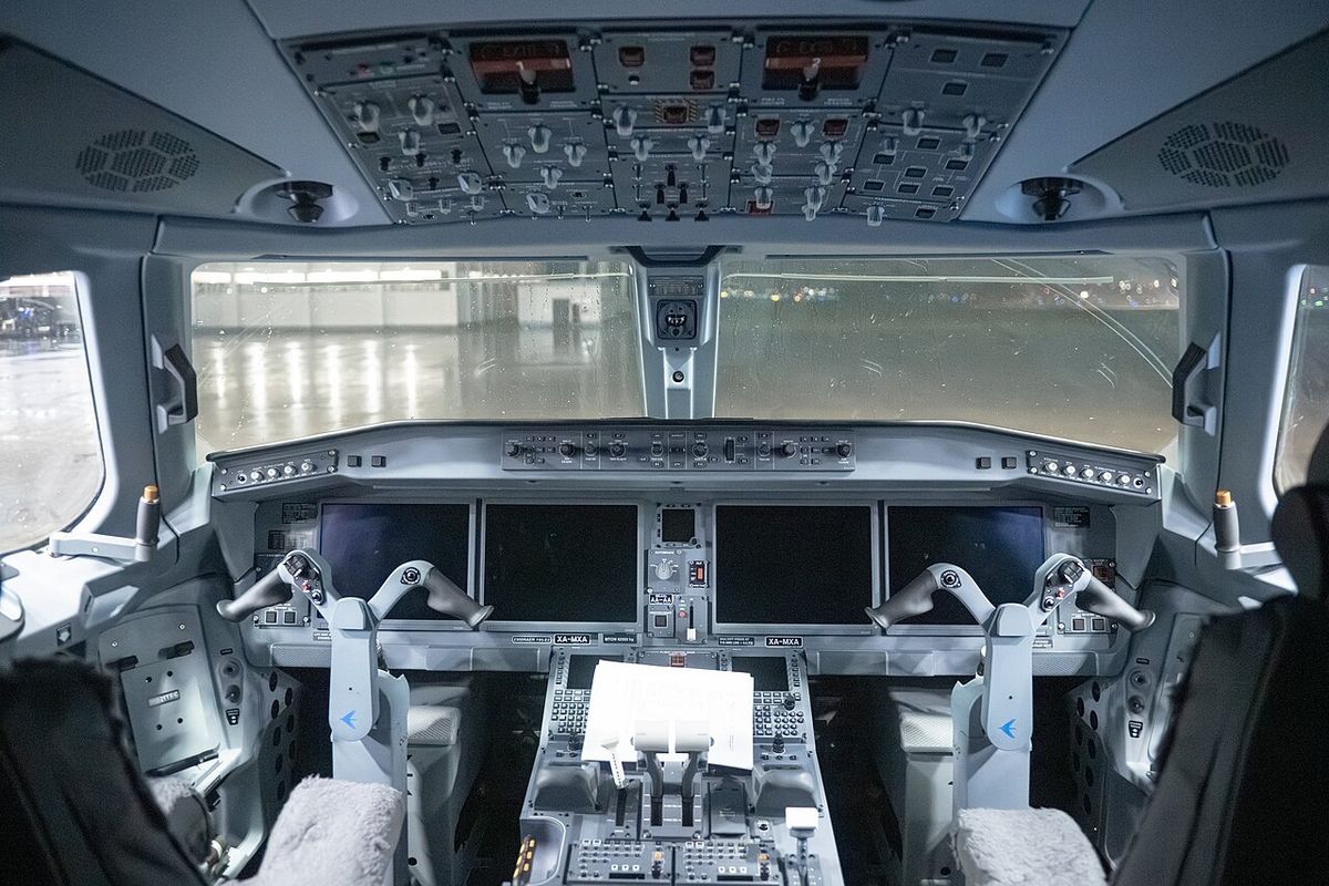 An airliner flight deck seen from behind the seats: four wide display screens across the panel, two control yokes, the pedestal and overhead panel.