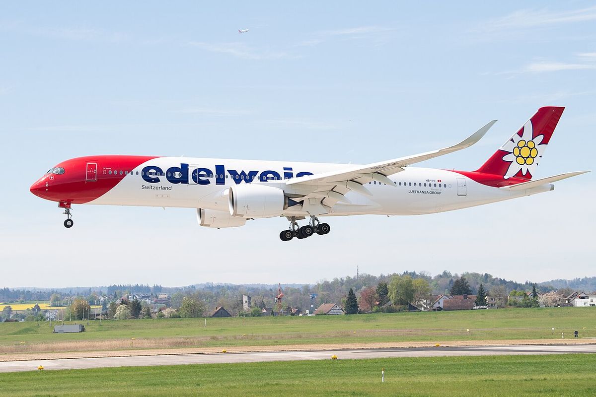 An Edelweiss Air Airbus A350 with a red nose and an edelweiss flower on the tail, landing with its gear down over green fields.