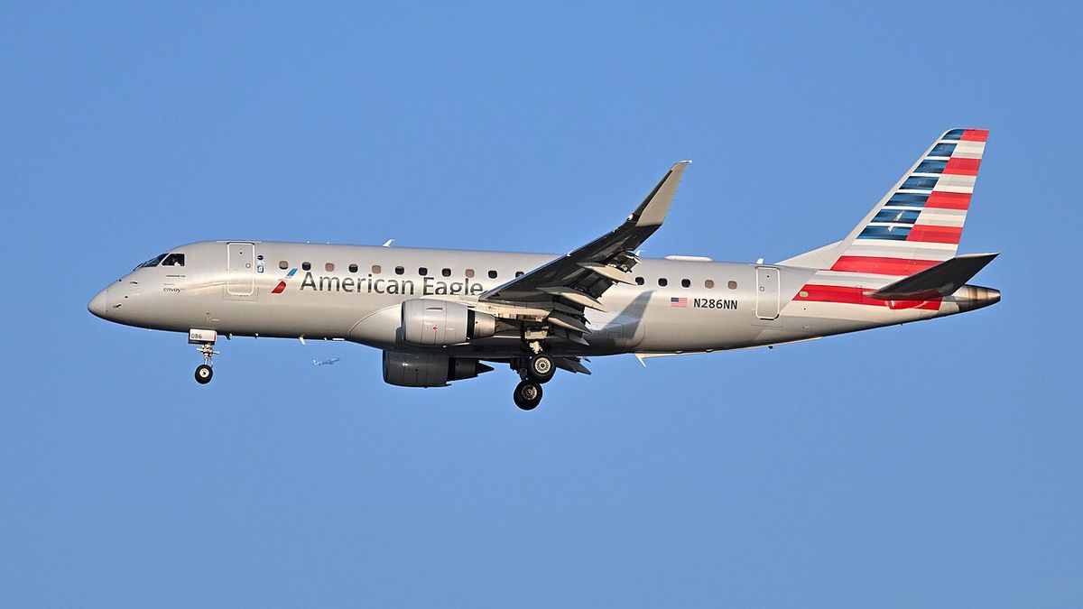 An American Eagle Embraer 175 in silver livery with the striped tail, gear down on final approach against a blue sky.