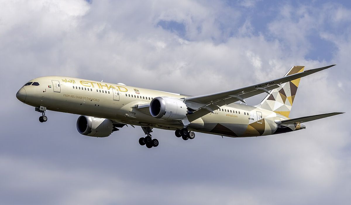 A beige and gold Etihad Boeing 787 with its landing gear down, flying under white and grey cloud.