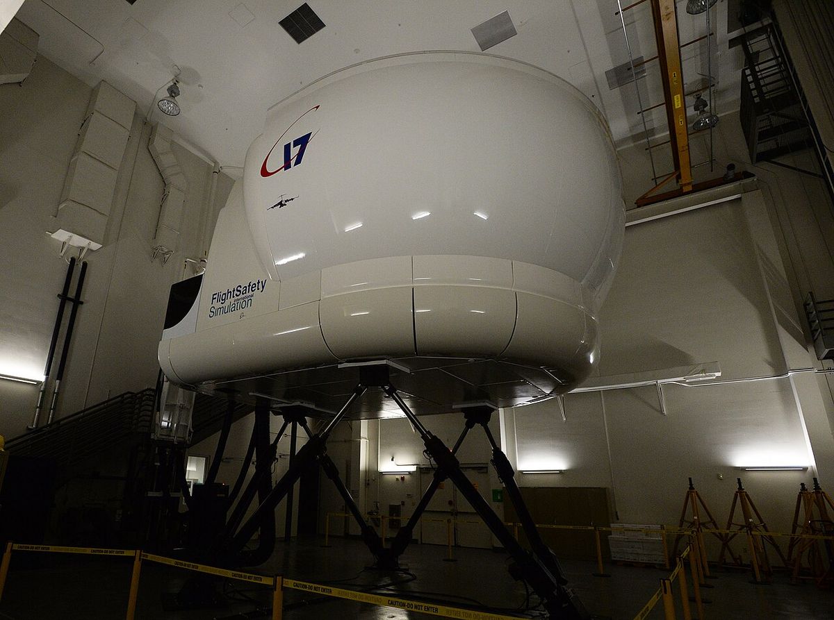 A large white enclosed simulator cab standing on long motion jacks in a dark hall, with a walkway at its rear.