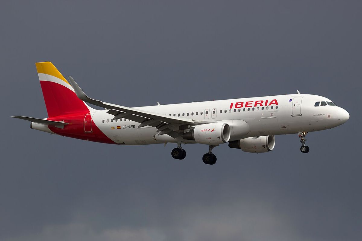 A white Iberia Airbus A320 with a red and yellow tail, landing gear down, in front of dark grey cloud.