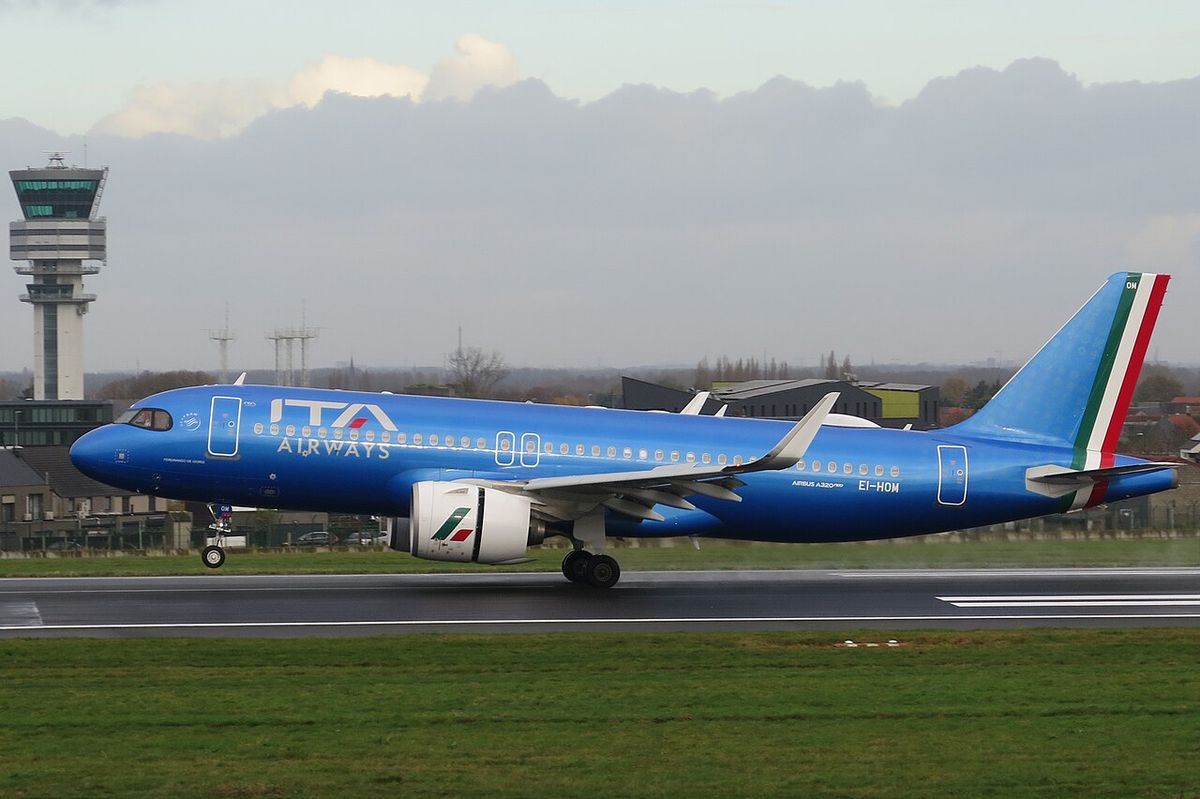 A bright blue ITA Airways Airbus A320neo with a tricolour tail edge touching down on a runway, a control tower behind.