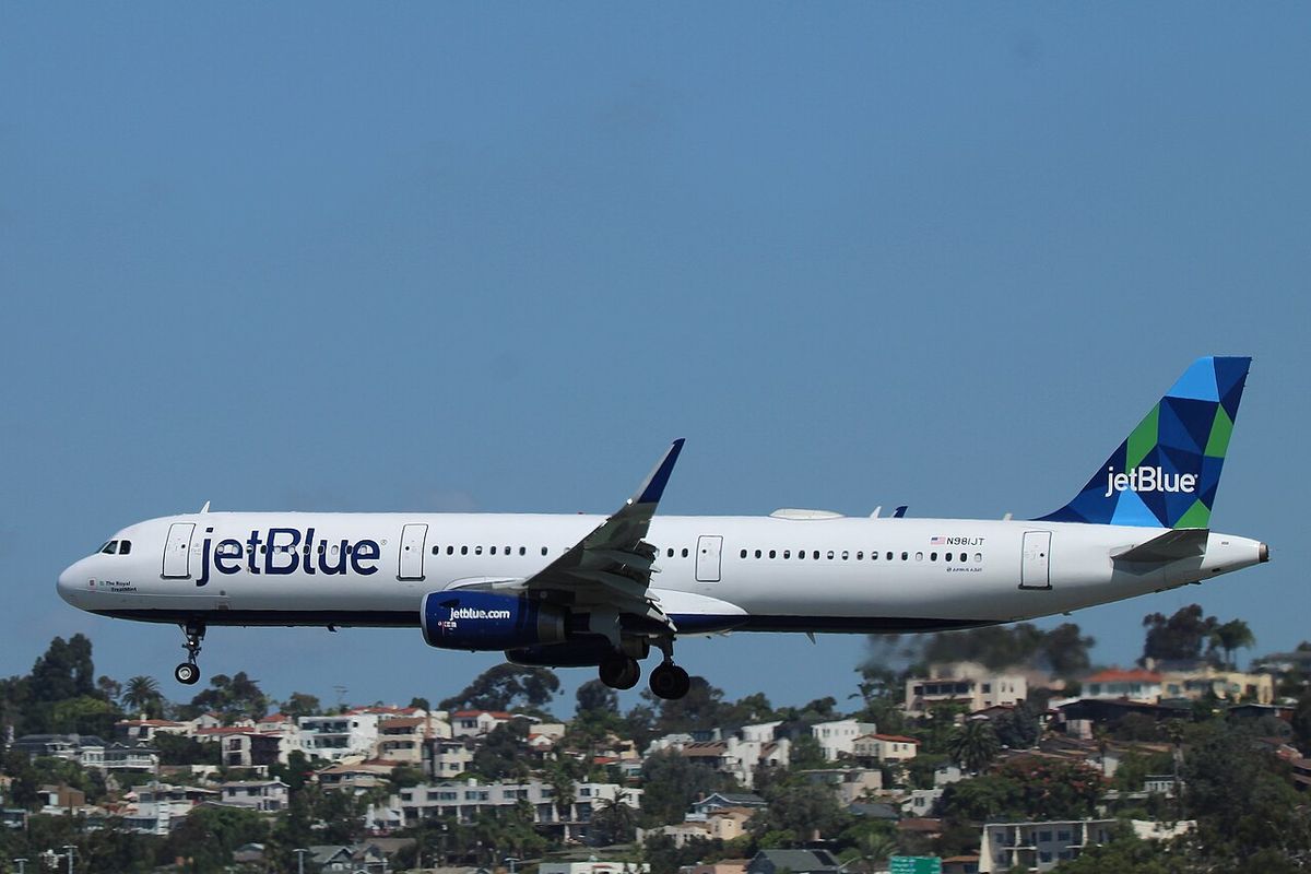 A white JetBlue Airbus A321 with a blue patterned tail flying low over hillside houses, landing gear down.