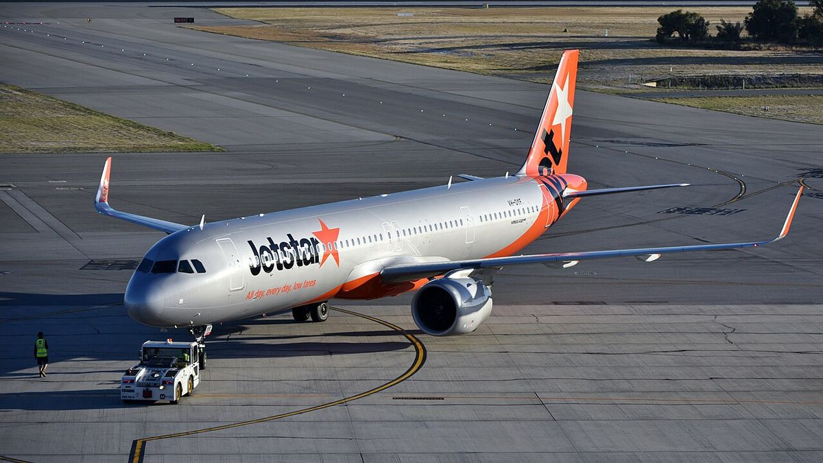 A Jetstar Airbus A321neo in silver livery with the orange star tail, being pushed back by a tug at an airport.