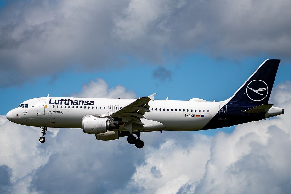 A white Lufthansa Airbus A320 with a dark blue tail and the crane emblem, landing gear down, below white and grey clouds.