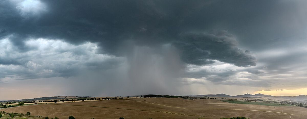 A dark storm cloud over open farmland, with a dense grey column of rain falling from it and fanning out at the ground.