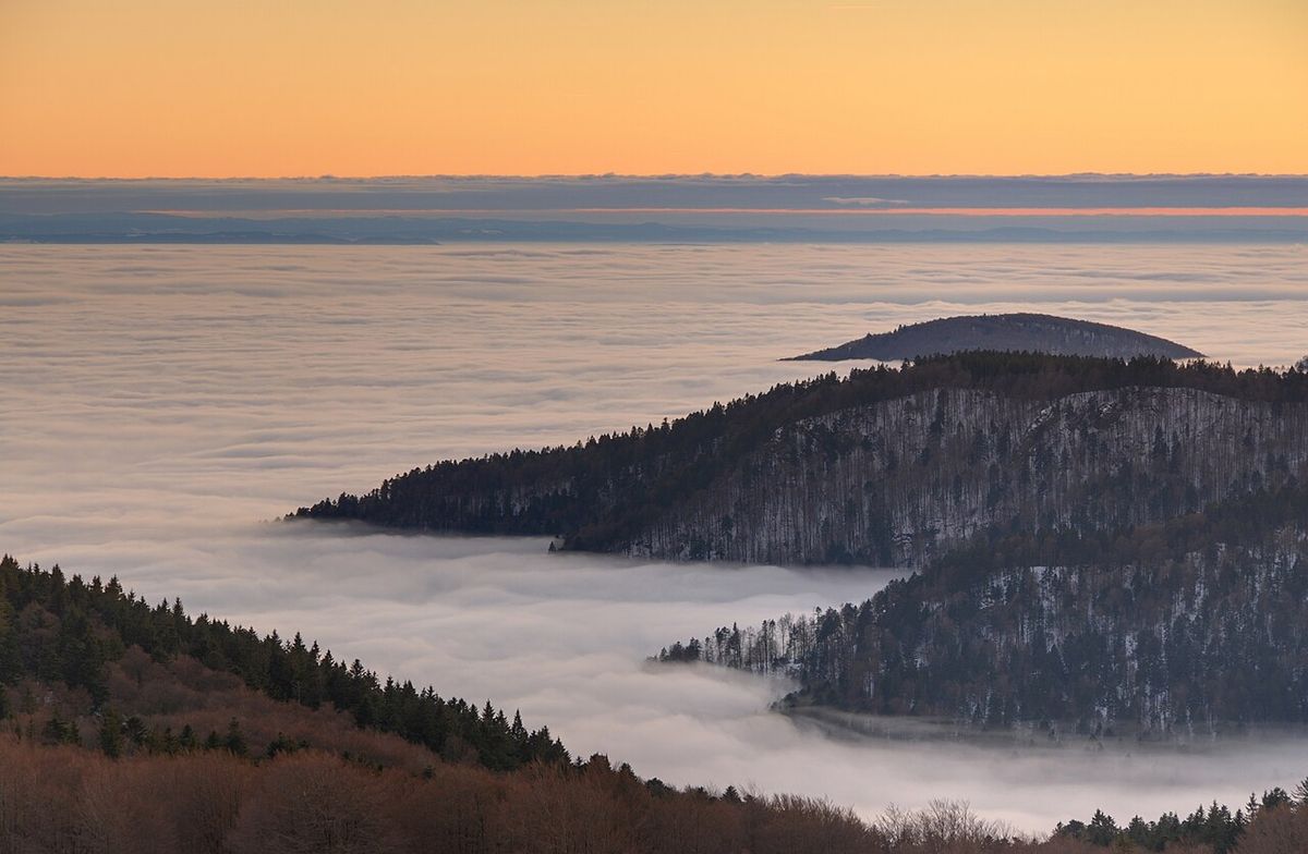 Seen from a mountain top at sunset, a flat white sea of fog fills the valley below, with dark forested ridges rising out of it.