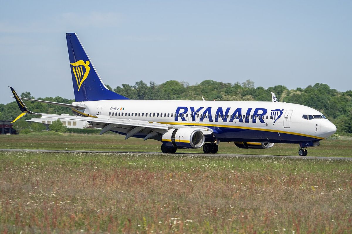 A white and blue Ryanair Boeing 737 with a yellow harp on its blue tail, rolling on a runway beside a strip of grass.