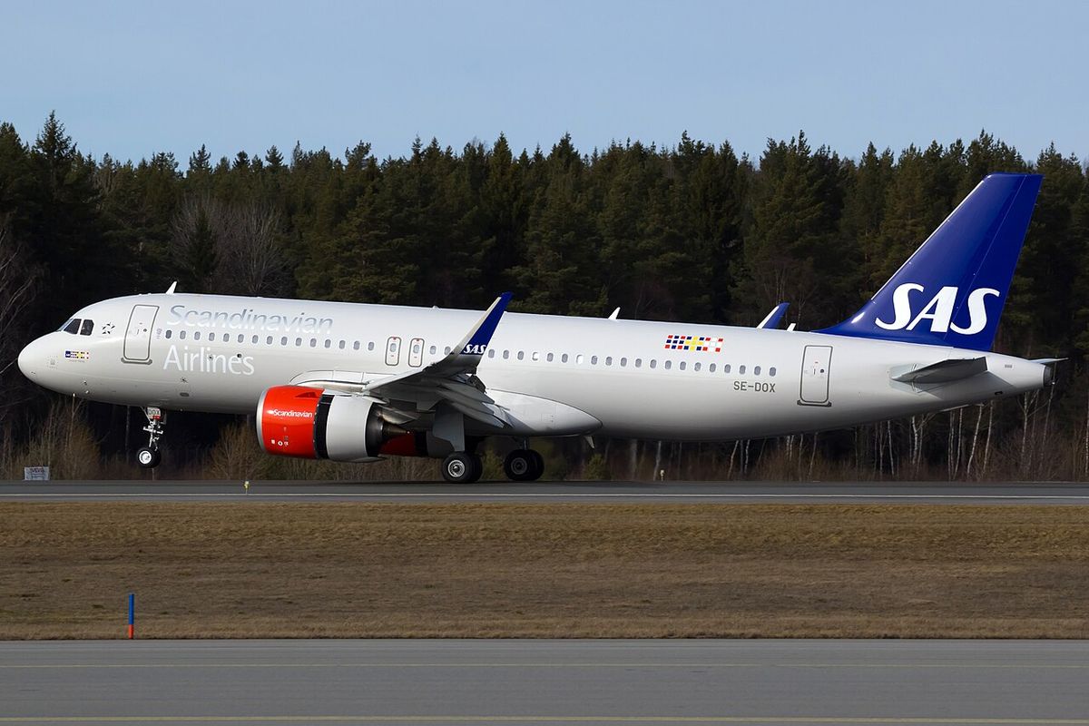 A white SAS Airbus A320neo with a dark blue tail and red engine cowls touching down on a runway, pine forest behind.