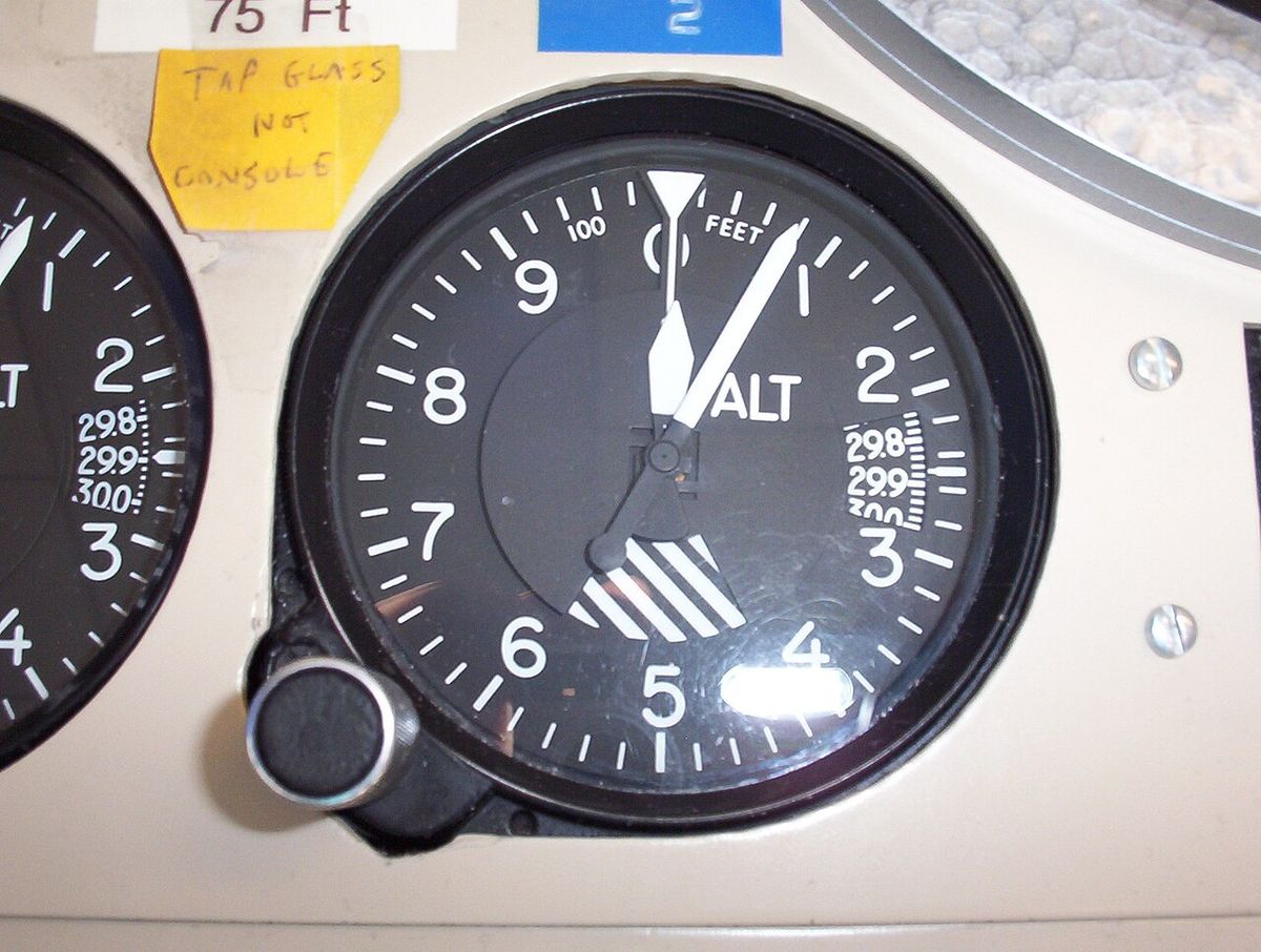 Round barometric altimeter reading about 75 ft, with pressure windows on the right and handwritten notes stuck on the console above it.