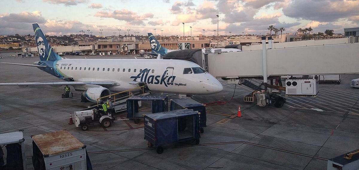 A white regional jet with a blue Alaska tail at a jet bridge at dusk, with baggage carts on the apron in front.