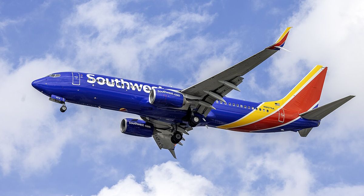 A dark blue Southwest Airlines Boeing 737 with a red and yellow tail, landing gear down, against blue sky and cloud.