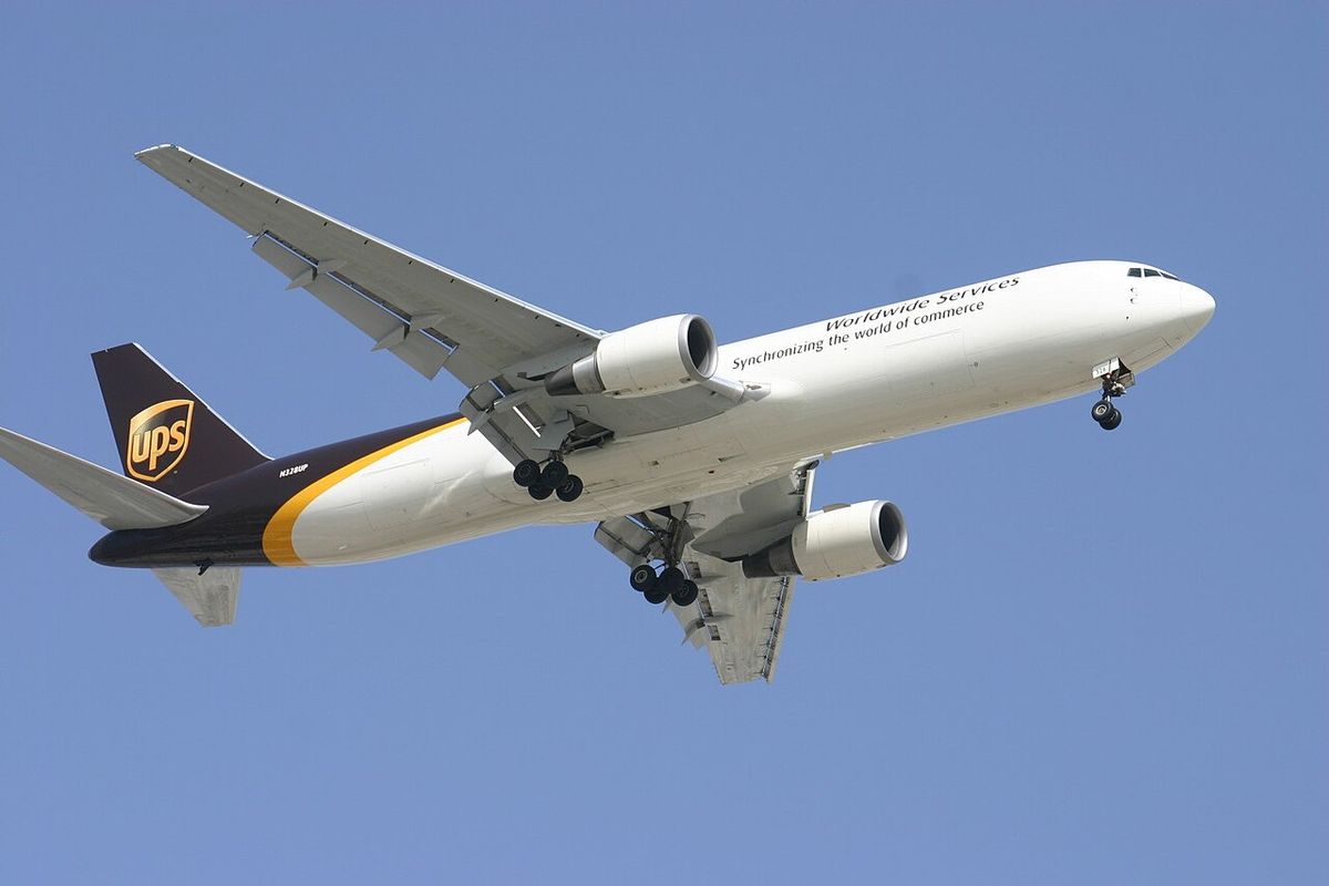 A UPS Boeing 767 freighter in brown, white and gold livery with its landing gear down, against a blue sky.