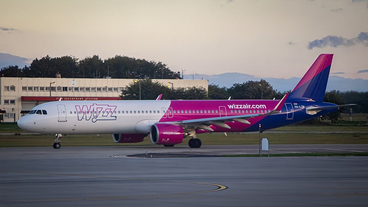 A long white Wizz Air airliner with a pink and purple tail and engines, on the ground beside a runway at dusk.